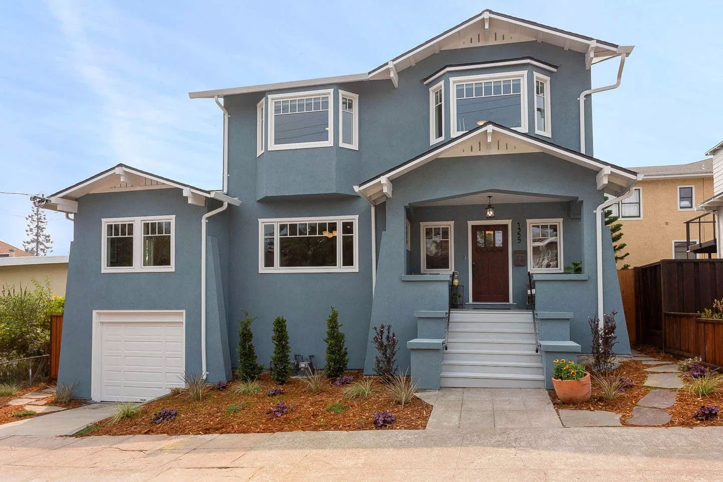 This is a photograph of a two-story residential house painted in light blue with white trim and a gray roof. The property features a garage on the left side, a front porch with steps leading up to it, and a well-maintained yard with a neatly trimmed lawn.