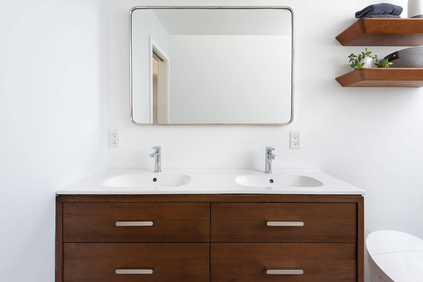 The image shows a modern bathroom with a double sink vanity, a large mirror above the vanity, wooden cabinets below the sinks, and a white wall with a painting hanging on it.
