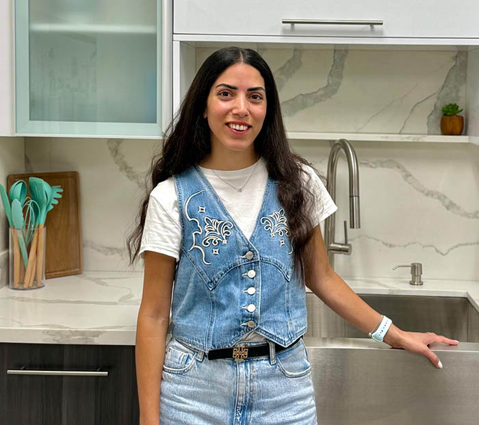 A woman with long hair is standing in front of a kitchen counter, smiling at the camera. She has on a denim vest over a white top, blue jeans, and a pair of sunglasses is placed on her head.