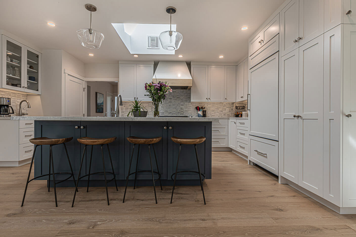 The image shows a modern kitchen interior with white cabinetry, a center island with bar stools, a dark countertop, and a large window allowing natural light into the space.