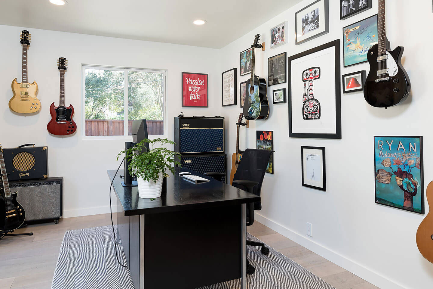 The image shows a room with walls covered in framed music memorabilia, guitars hanging on the wall, musical equipment such as amplifiers and microphones, and various other decorative items related to music and musicians. There is a desk with a computer monitor, keyboard, and chair, suggesting a workspace or home office setting.