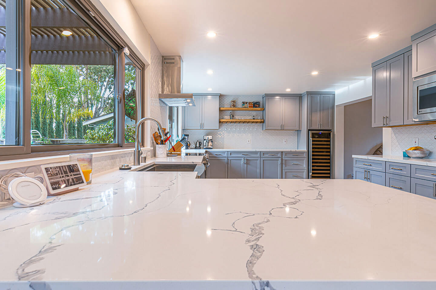 The image shows a modern kitchen interior with a large island featuring a marble countertop, cabinets with glass doors, and a backsplash. The kitchen has stainless steel appliances, a sink, and a window with natural light.